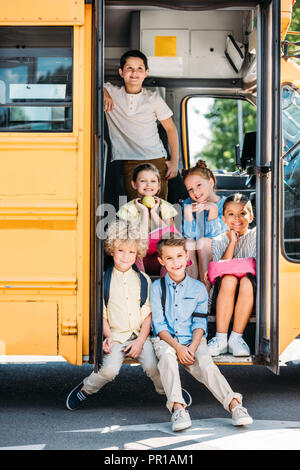 Groupe d'écoliers mignon assis sur les escaliers de l'autobus scolaire et looking at camera Banque D'Images