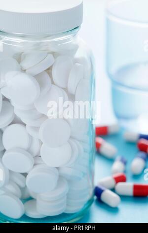 Pilules blanches dans une bouteille en verre et un verre d'eau avec divers autres capsules de drogue dans l'arrière-plan. Banque D'Images