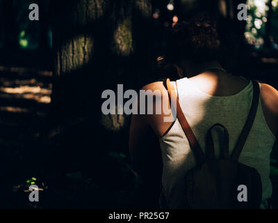 Une fille noire à l'arrière avec un sac à dos sac à dos avec sa tête vers le bas en face d'un arbre avec des rayons du soleil à travers feuilles très douillet, la peur Banque D'Images