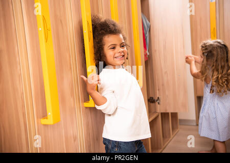 Les enfants multiculturels souriant debout près des casiers dans la maternelle vestiaire Banque D'Images