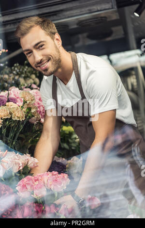 Voir à travers la vitre du fleuriste beau sourire en prenant soin de bouquets de fleurs et looking at camera Banque D'Images