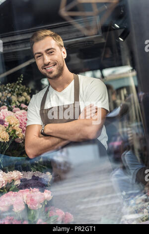 Voir à travers la vitre du beau fleuriste à tablier, debout les bras croisés dans le magasin de fleurs et looking at camera Banque D'Images