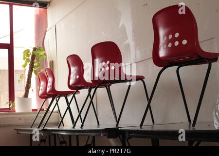 Des chaises en plastique moulé rouge debout sur des tables pliantes avec une fenêtre rouge en arrière-plan et de murs blancs. Banque D'Images