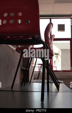 Des chaises en plastique moulé rouge debout sur des tables pliantes avec une fenêtre rouge en arrière-plan et de murs blancs. Banque D'Images