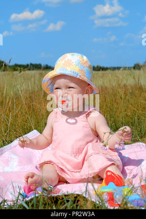 Cute little girl in a hat assis sur une couverture rose, vert et bleu ciel sur le terrain à l'arrière-plan Banque D'Images