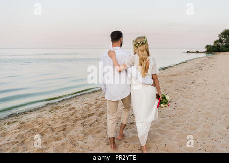 Vue arrière de la mariée avec mariage bouquet et groom hugging and walking on beach Banque D'Images