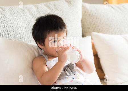 Little asian girl main tenant le verre de lait de consommation, assis sur un canapé à la maison. La médecine et les soins de concept. Banque D'Images