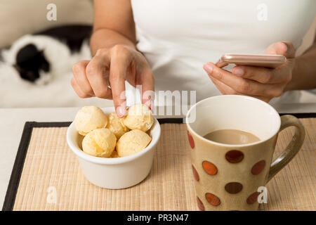 Close up of Woman picking ball de pain fromage (connu sous le nom de Pao de Queijo" au Brésil) et holding cell phone dans l'autre main. Tasse de café et comestic Banque D'Images