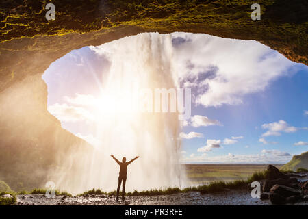 Happy female traveler sentir la puissance de la cascade de Seljalandsfoss dans le sud de l'Islande, personne debout derrière le cours d'eau, coucher de la lumière, intense Banque D'Images