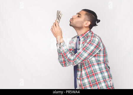 Vue latérale du profil cheerful riche beau jeune homme élégant barbu en chemise à carreaux colorés et de l'air permanent kissing fan de dollars. Ind Banque D'Images