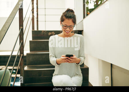 Young Asian businesswoman wearing glasses and smiling while sitting on stairs dans un bureau à l'aide d'un téléphone cellulaire pendant sa pause Banque D'Images