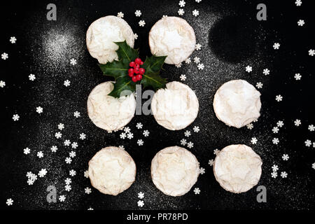 Petits pâtés de Noël avec un manque, baie de houx sprig, feuilles et saupoudre de sucre glace flocon de poussière sur fond noir. Mise à plat. Banque D'Images