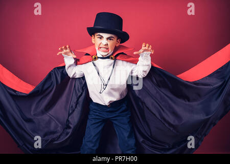 Halloween vampire boy agite sa cape rouge, noir avec ses mains entouré sur red studio arrière-plan. Maquillage de vampire pour enfants. Banque D'Images