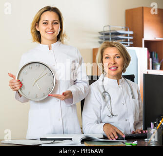 Deux femelles en combinaisons blanches holding clock and smiling in clinique moderne Banque D'Images