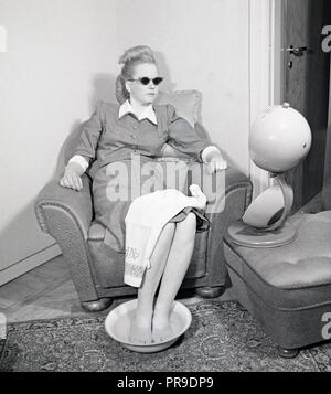 Soins de beauté en 1940s. Une jeune femme se détend dans un fauteuil confortable tout en bronzant et en faisant un bain de pieds. La lumière artificielle du soleil des équipements de bronzage à la maison comme cette lampe solaire a donné un bronzage et a parfois été un traitement pour des maladies. Vous devez porter des lunettes de soleil de protection lorsque vous les utilisez. Suède 1947. Photo Kristoffersson AB22-1 Banque D'Images