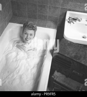 1940 femme dans la baignoire. Une jeune femme est d'avoir un bon bain chaud. Elle fait attention de ne pas obtenir de ses cheveux mouillés, et il a sous un foulard. Une radio est debout sur une chaise à côté de la baignoire, et elle est à son écoute tout en se baignant. Sur le panneau avant de la radio Vous pouvez choisir à l'aide d'une molette démarré. Suède 1945. Kristoffersson Photo ref L104-4.