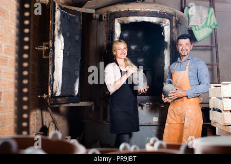 Smiling man and woman holding potiers de vases en céramique émaillée noire à côté du four Banque D'Images