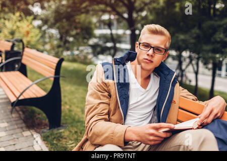 Sac à dos College student with reading book walking in autumn park assis sur un banc. Man studying outdoors Banque D'Images