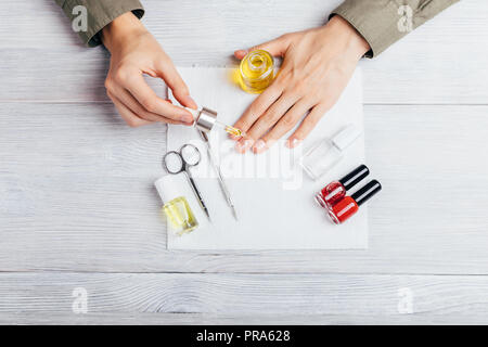 Femme appliquer nourrir d'huile sur les cuticules des instruments pour manucure, bouteilles de vernis rouge et de base, vue du dessus sur le tableau blanc. Jeune femme soins o Banque D'Images