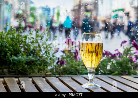 Verre de bière blonde sur une table en bois et de gouttes de pluie sur la fenêtre humide, floue méconnaissable les gens marchent sur la route. Notion de temps de pluie, saisons, ville moderne Banque D'Images