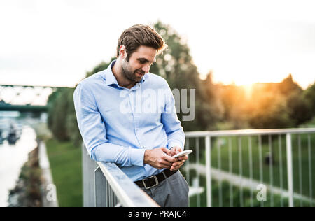 Un jeune homme avec le smartphone debout sur un pont au coucher du soleil, s'appuyant sur une balustrade. Banque D'Images