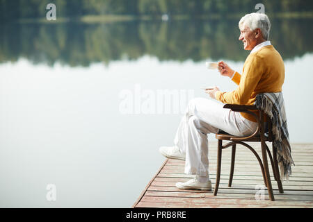 Happy handsome man détendue portant des vêtements élégants assis sur la vieille chaise avec couverture sur Pier et de boire du café tout en contemplant le lac Banque D'Images