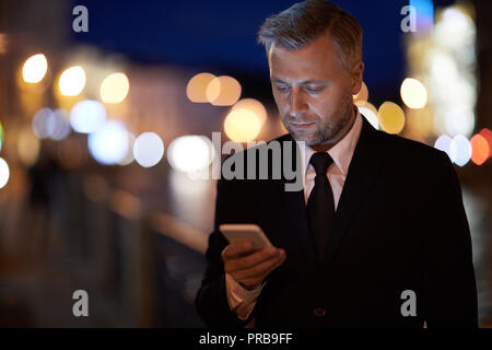 Homme sérieux en soirée textos en smartphone dans l'environnement urbain de nuit Banque D'Images