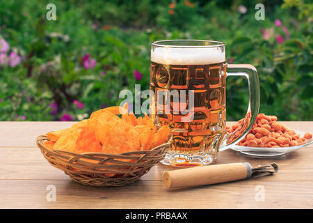 Tasse en verre de bière sur la table en bois avec des croustilles dans panier en osier, les arachides dans la plaque, sur fond vert naturel de l'ouvreur Banque D'Images