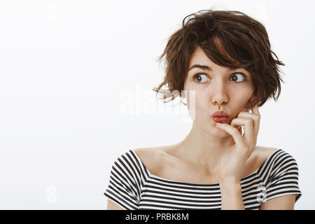 Tourné à l'intérieur de curieux emotive libre de cute hairstyle malpropre, le pliage des lèvres et dire wow tout en tenant la main près de la bouche, à gauche avec intérêt et curiosité sur mur gris Banque D'Images