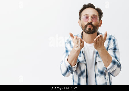 Charmant homme gay adultes satisfait avec l'expression heureuse du quartier branché de lunettes de soleil et serra les lèvres de pliage, chemise et souriant tout en envoyant des baisers à l'appareil photo, maintenant près de palmiers donnant bouche mwah Banque D'Images