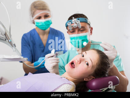 Portrait de peur teen girl avec la bouche ouverte pendant un traitement dentaire avec deux dentistes debout près de présidence Banque D'Images