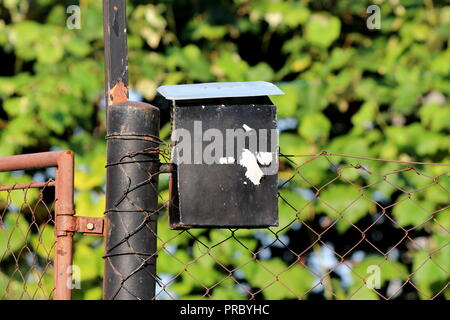 Fortement utilisée vieille petite black metal avec boîte aux lettres délabrées et peinture craquelée partiellement monté sur poteau rouillé avec grillage attaché à droite Banque D'Images