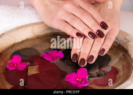 Jeune fille brune avec des mains gel manucure polonais sur les doigts au-dessus de l'eau avec des fleurs violettes et les pierres noires en bol en bois en studio. Close up Banque D'Images