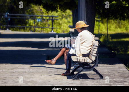 Elder African American Woman est assis sur le banc en bois dans un parc et navigue sur son smartphone. Banque D'Images