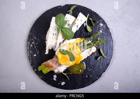 Pain grillé avec des bâtons d'asperges vertes grillées enrobé de bacon, oeuf poché , pousses et de fromage bleu sur fond ardoise noire. Gray conc Banque D'Images