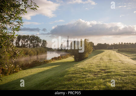 Un paysage pittoresque d'un moulin à vent hollandais traditionnel le long d'une rivière au début de matin brumeux en Hollande. Banque D'Images