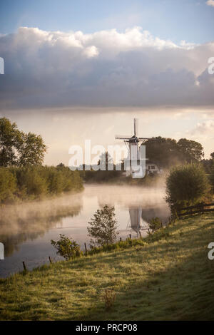 Un paysage pittoresque d'un moulin à vent hollandais traditionnel le long d'une rivière au début de matin brumeux en Hollande. Banque D'Images