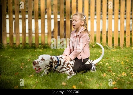 Souriant, chantant little girl hugging un gros chien dans le pré,jeux de plein air.Heureux l'enfant aime son animal de compagnie. Photo de Positive little girl hugging adorable dalmatien, portant sur l'herbe.chien Dalmatien. Banque D'Images