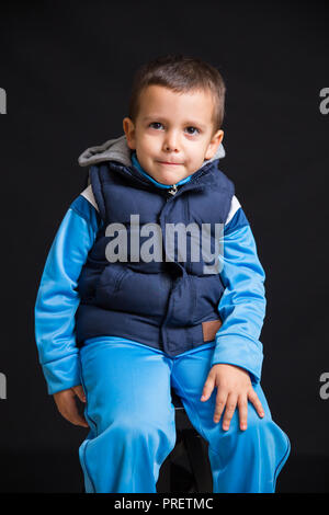 Portrait de penser petit garçon assis sur un tabouret noir, isolé sur fond noir Banque D'Images