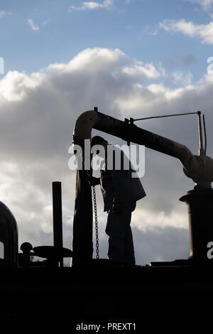 Gros plan de l'équipage du train à vapeur ferroviaire, en silhouette de ciel, debout sur la locomotive britannique vintage à l'arrêt de l'eau, prise de moteur sur l'eau, en début de soirée. Banque D'Images