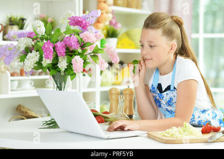 Cute girl using laptop durant la préparation d'une salade fraîche sur la table de cuisine Banque D'Images