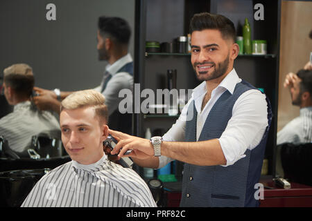 Beau et confiant de coiffure cheveux du client de fraisage avec massicot. Coiffure barbu en chemise blanche et gilet looking at camera et souriant. Jeune homme en robe coupe à la route. Banque D'Images