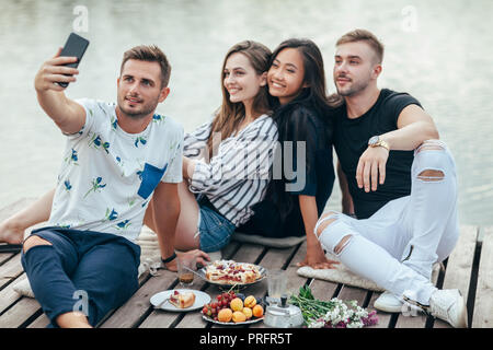 Amis sitting on pier selfies avec lake arrière-plan tout en se reposant à pique-nique. Amitié et détente concept Banque D'Images