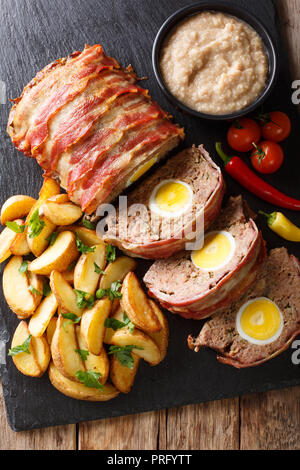 Pain de viande fraîchement préparé avec des œufs enveloppés dans le lard avec les quartiers de pommes de terre et légumes gros plan sur un fond noir. Haut Vertical Vue de dessus Banque D'Images