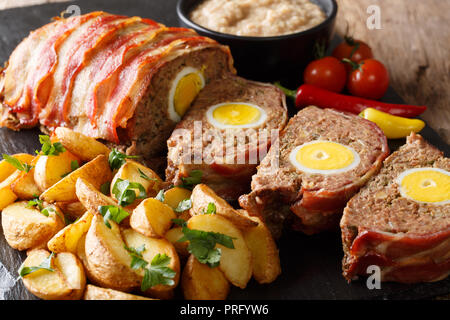 Pain de viande fraîchement préparé avec des œufs enveloppés dans le lard avec les quartiers de pommes de terre et légumes gros plan sur un fond noir. Banque D'Images