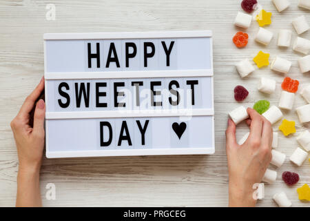 Conseil moderne avec texte 'Happy Sweetest Day' mot et femmes avec des bonbons sur fond de bois blanc, vue du dessus. À partir de ci-dessus, mise à plat, les frais généraux. Banque D'Images