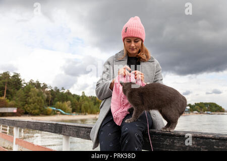 Close-up of woman knitting mains chapeau en laine rose avec aiguille, next est un beau chat gris contre le fond de la mer dans la journée ensoleillée d'automne Banque D'Images