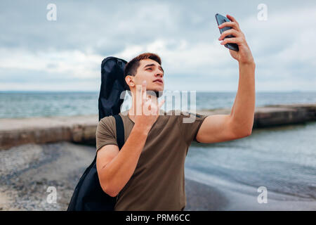 Jeune homme portant la guitare acoustique et de prendre l'aide de selfies téléphone sur nuageux beach. Guy montre rockstar symbole avec la main. Banque D'Images