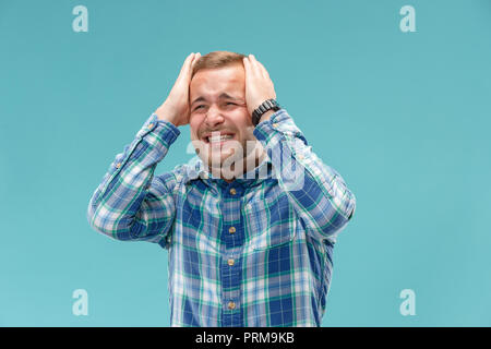 Horrible, stress, choc. Portrait de mi-longueur mâle isolée au studio bleu. Jeune homme malheureux émotionnelle qui serre la tête dans les mains. Les émotions humaines, l'expression faciale concept. Couleurs tendance Banque D'Images