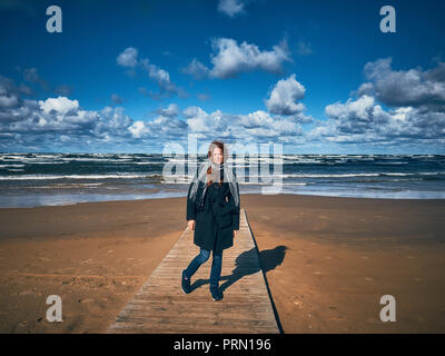 Jeune fille rousse vêtue d'un manteau noir promenades le long du littoral. Un fort vent a attisé les boucles de la jeune fille. Banque D'Images